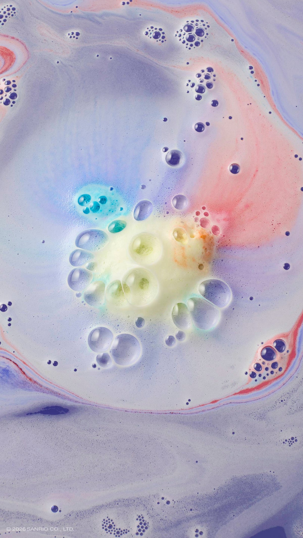 An aerial shot of lavender bath water swirling with multi-coloured foam. In the centre, a cluster of yellow and blue bubbles marks where the "U.SA.HA.NA" bath bomb has dissolved, with soft streaks of pink and purple radiating across the surface.